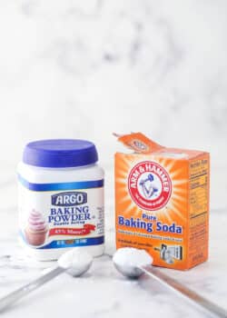 a container of baking soda and baking powder sitting on marble background with a scoop of each in front