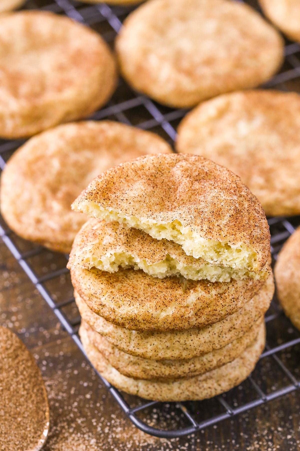Snickerdoodles are made of buttery sugar cookie dough rolled into balls, coated in cinnamon sugar, and baked into chewy, tangy cookies. A stack of snickerdoodles on a wire rack. The top cookie has a bite taken out of it.