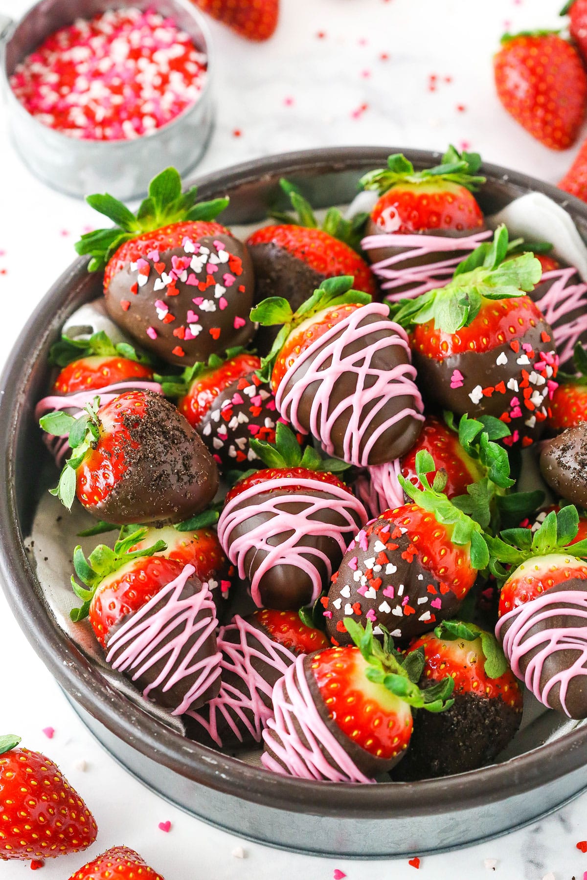 Chocolate Covered Strawberries may look fancy, but they are so easy to make! Learn how to make & decorate chocolate covered strawberries. Chocolate Covered Strawberries decorated with pink frosting or red, white and pink sprinkles stacked in a glass bowl on a white table top