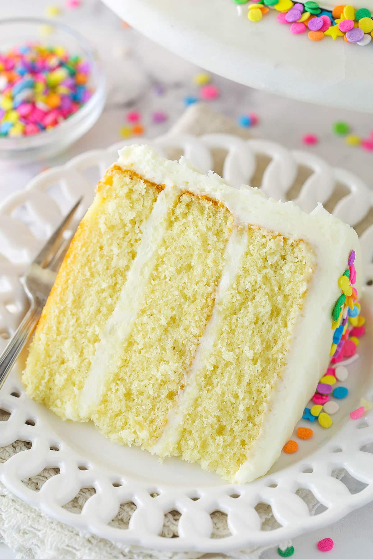 This moist vanilla layer cake is made of three layers of moist vanilla cake that are filled and frosted with rich vanilla buttercream. A slice of moist vanilla layer cake on a plate with a fork.