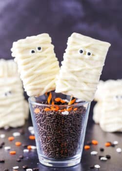 Two Mummy Rice Krispie Treats on orange and black straws in a glass filled with chocolate sprinkles.
