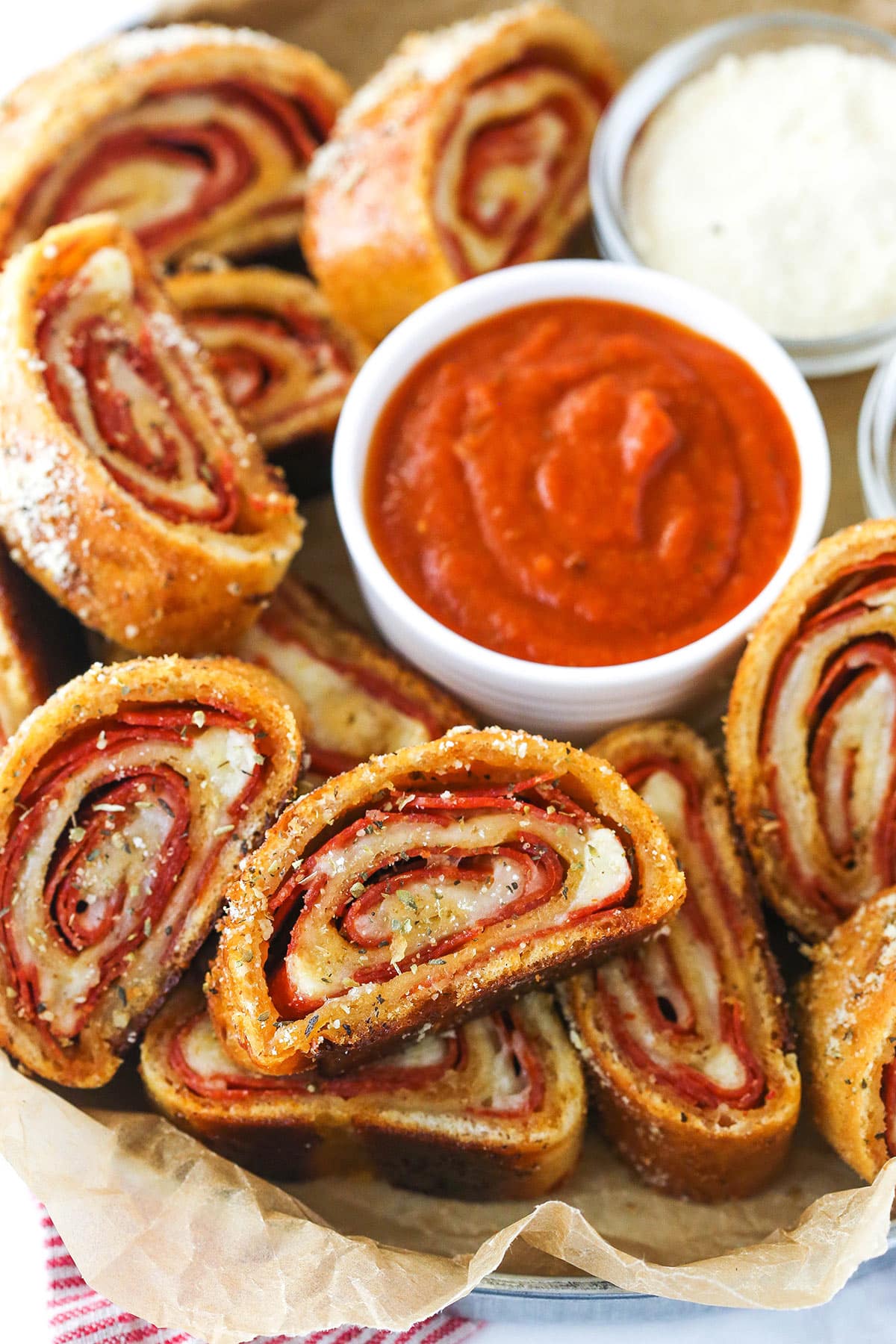 Pepperoni rolls are made up of pizza dough brushed with butter, wrapped around pepperoni and mozzarella cheese, and baked to golden brown. Closeup of pepperoni rolls with marinara sauce and parmesan cheese on a serving tray lined with parchment paper.