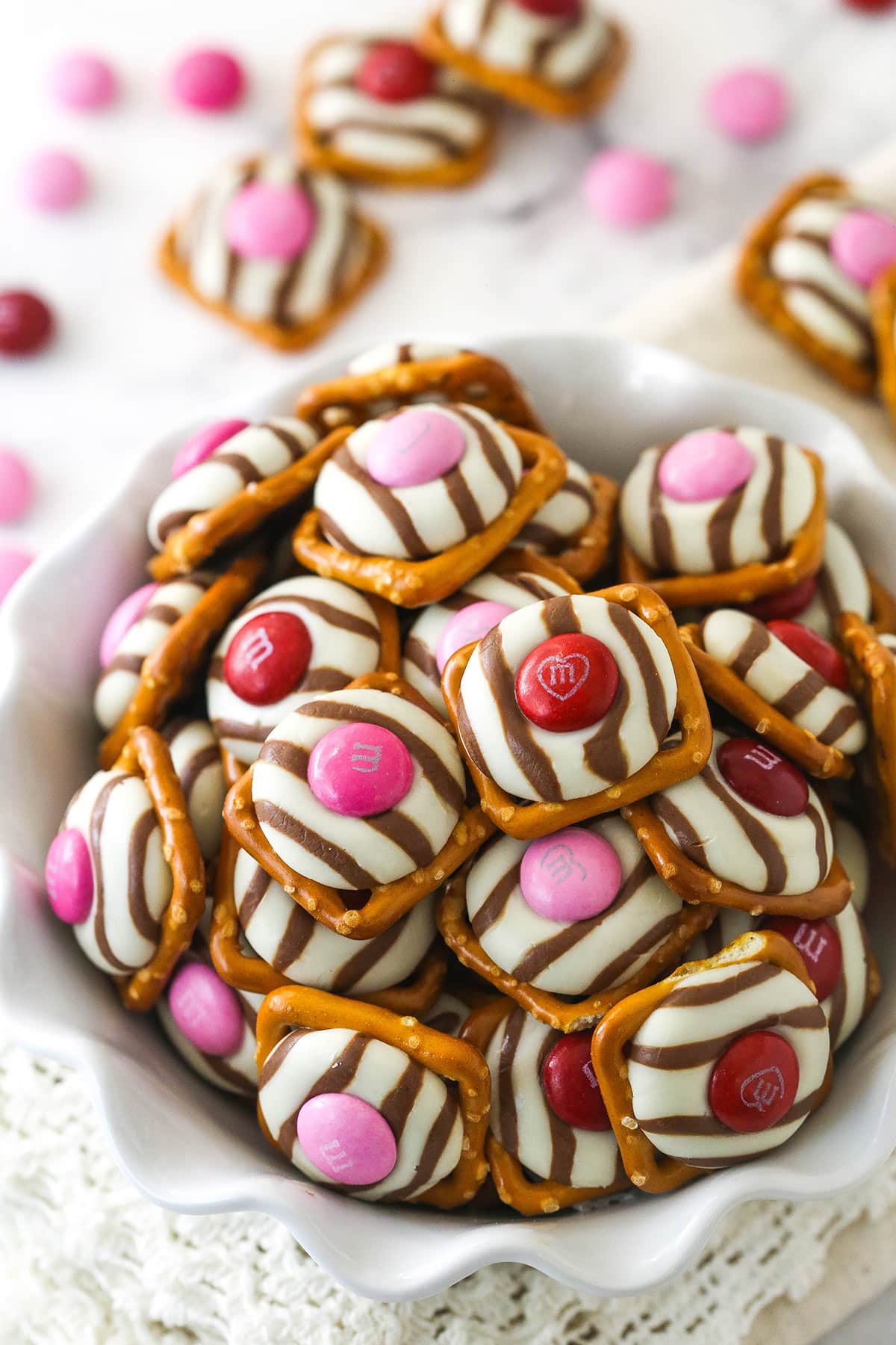 These Pretzel M&M Hugs are festive little chocolate treats! They are easy to make, use only 3 ingredients and you can easily change up the colors for any holiday! pink and red valentine's day pretzel M&M hugs in a white ruffle bowl