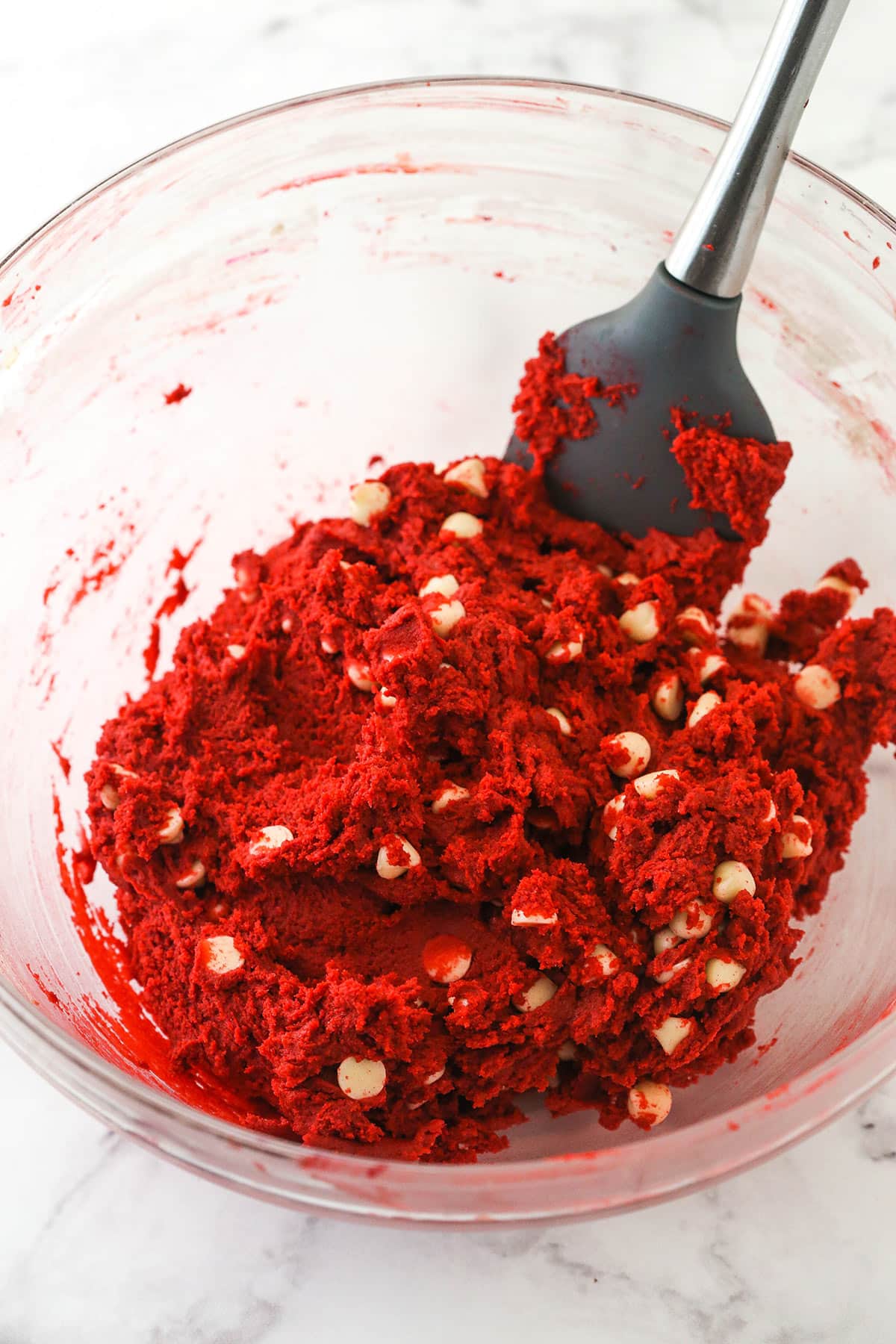 A spatula folding white chocolate chips into a bowl of red velvet cookie dough.