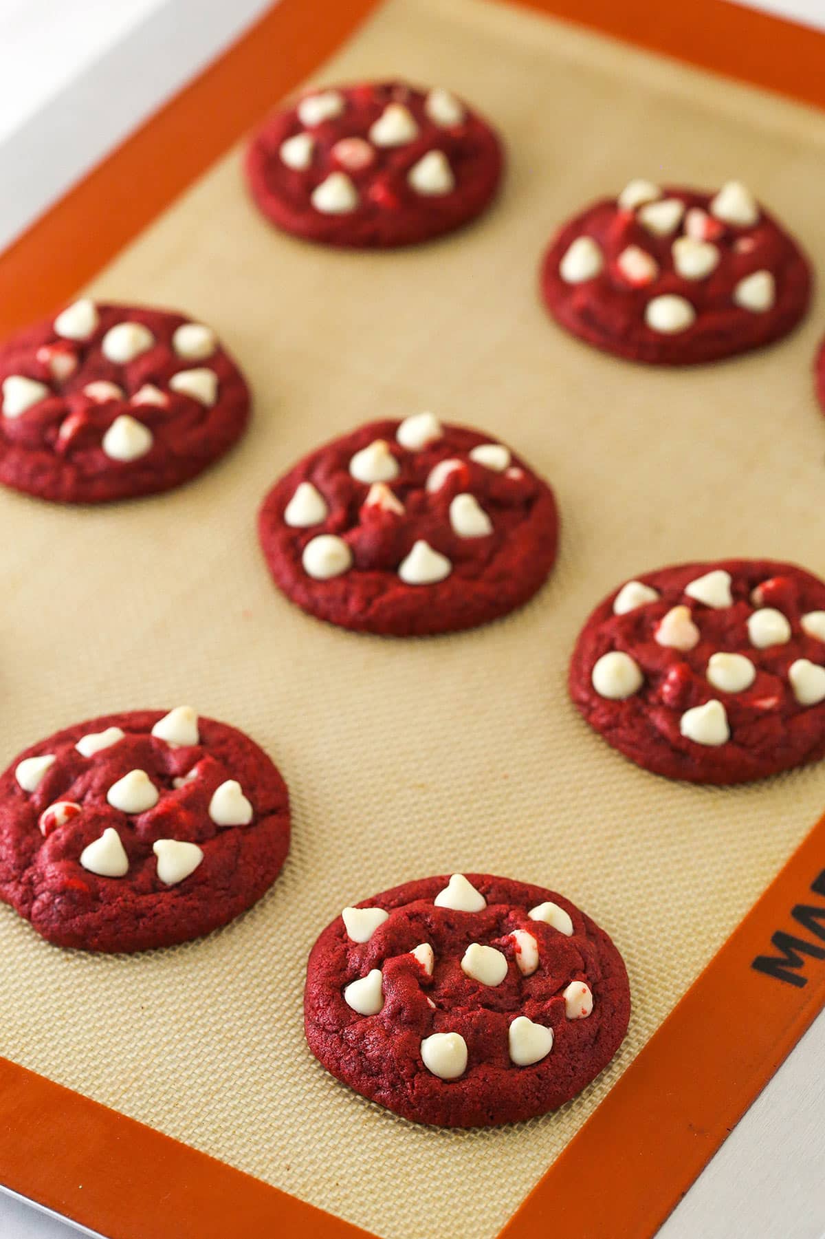 Baked red velvet cookies with white chocolate chips on a cookie sheet lined with a silicone baking mat.
