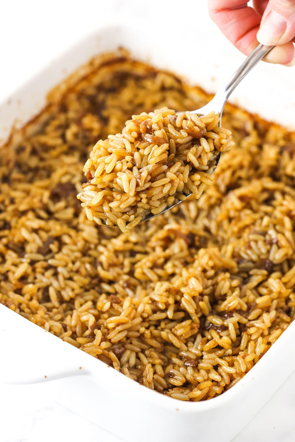 A metal spoon holding up a big bite of baked rice with a pan full of more rice beneath it