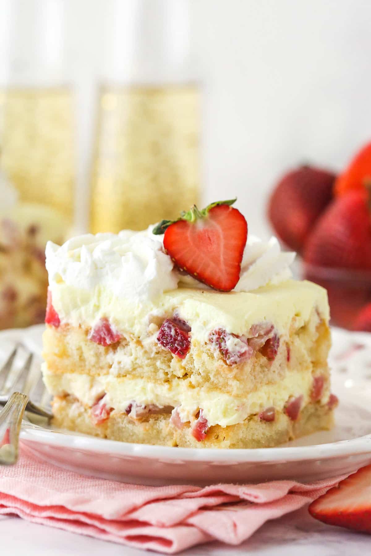 Strawberry Champagne Tiramisu is such a fun and tasty twist on classic tiramisu. Ladyfingers are dipped in a lightly-sweet champagne mixture and layered with chopped strawberries and delicious mascarpone filling. A slice of Strawberry Champagne Tiramisu on a white plate and pink napkin.