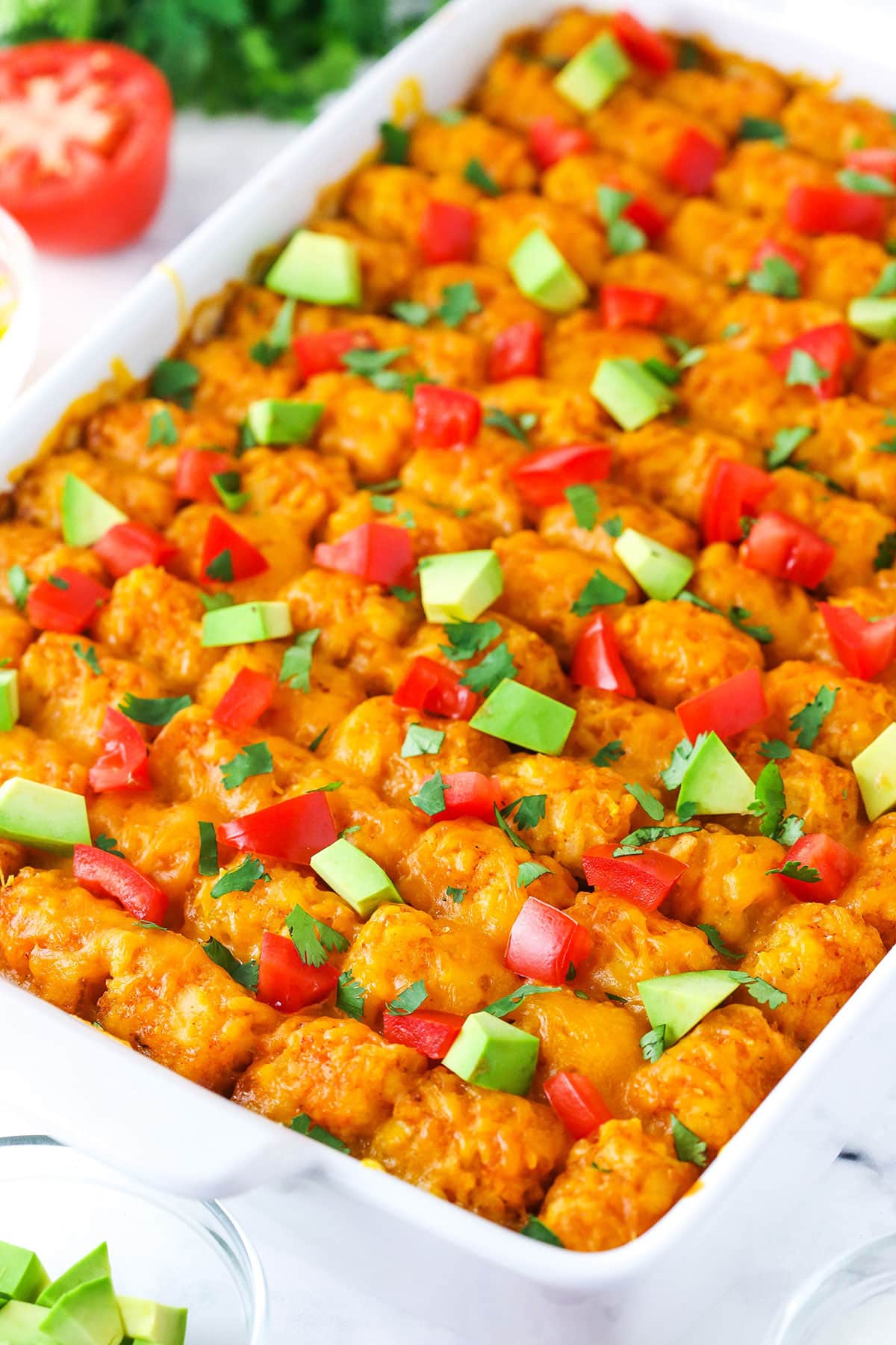 Taco tater tot casserole combines crispy tater tots and your favorite taco ingredients! This cheesy tater tot hotdish is so good. Taco Tater Tot Casserole with chopped avocado and tomato on top in a white platter