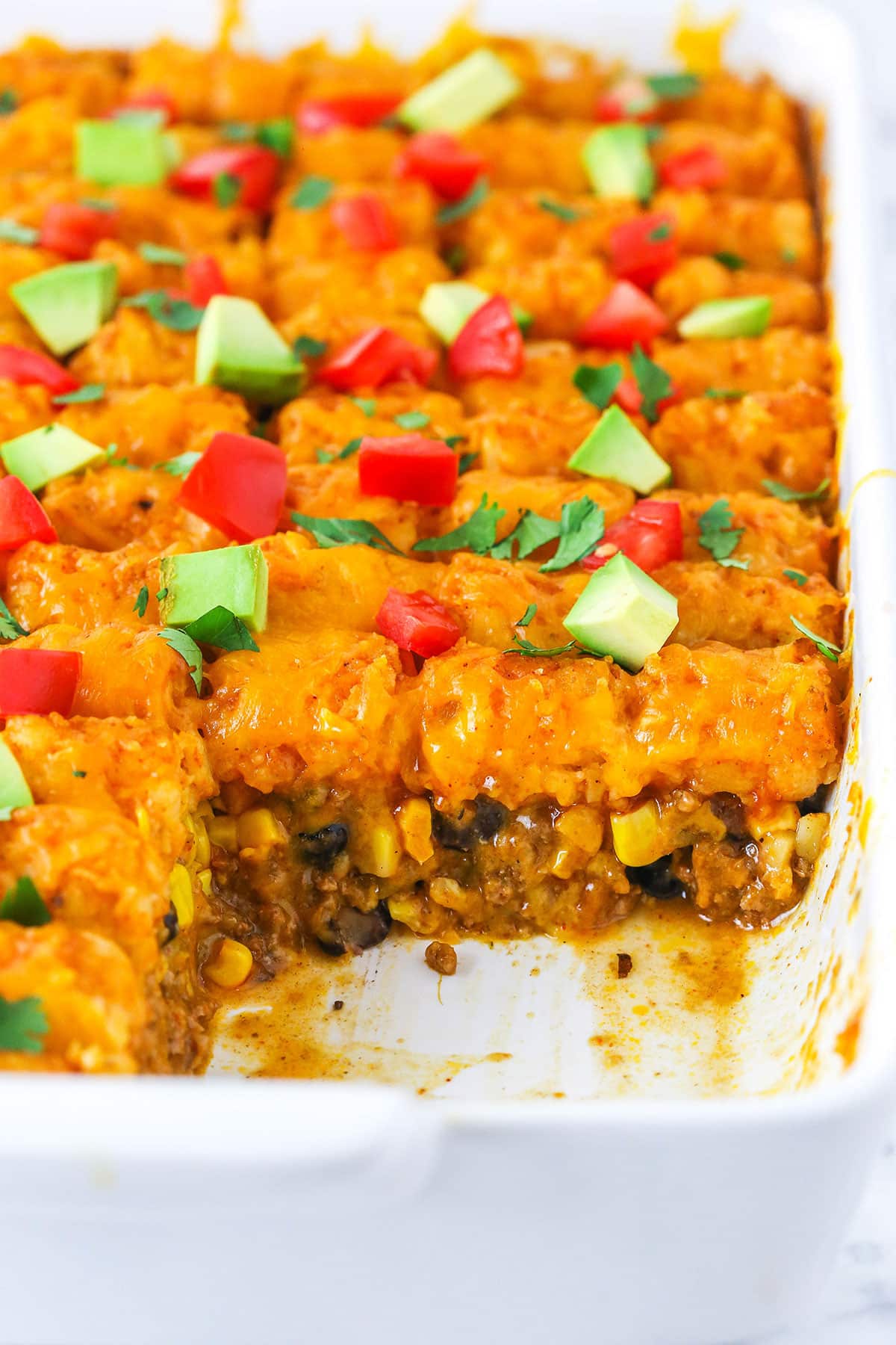 Taco tater tot casserole combines crispy tater tots and your favorite taco ingredients! This cheesy tater tot hotdish is so good. Taco Tater Tot Casserole with a serving removed in a white platter