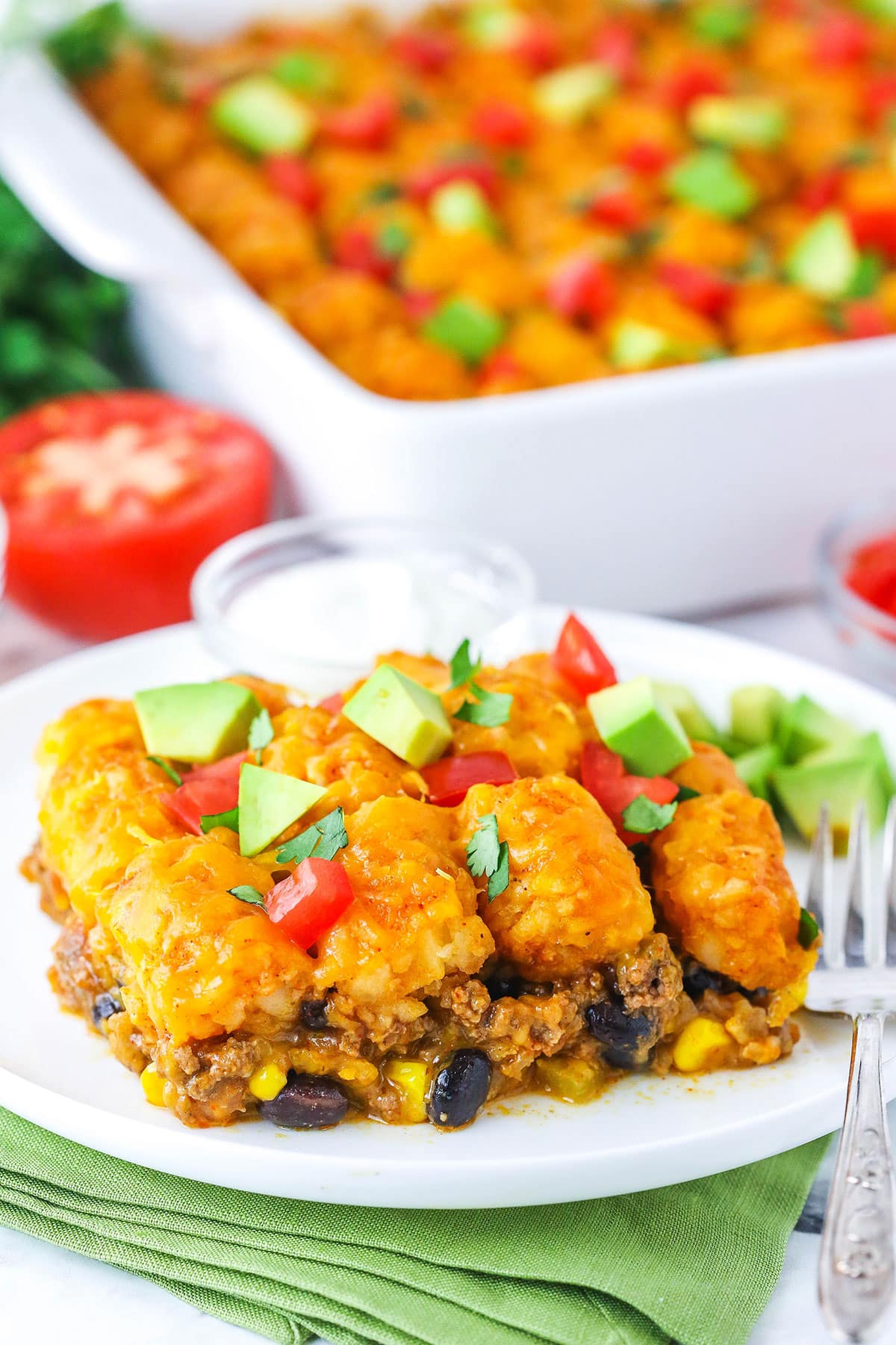 Taco tater tot casserole combines crispy tater tots and your favorite taco ingredients! This cheesy tater tot hotdish is so good. A serving of Taco Tater Tot Casserole topped with chopped avocado and tomato on a white plate with a fork