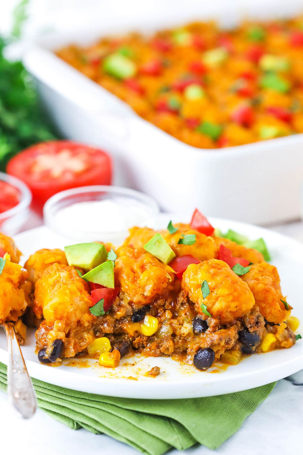 Taco tater tot casserole combines crispy tater tots and your favorite taco ingredients! This cheesy tater tot hotdish is so good. A serving of Taco Tater Tot Casserole topped with chopped avocado and tomato on a white plate with a fork