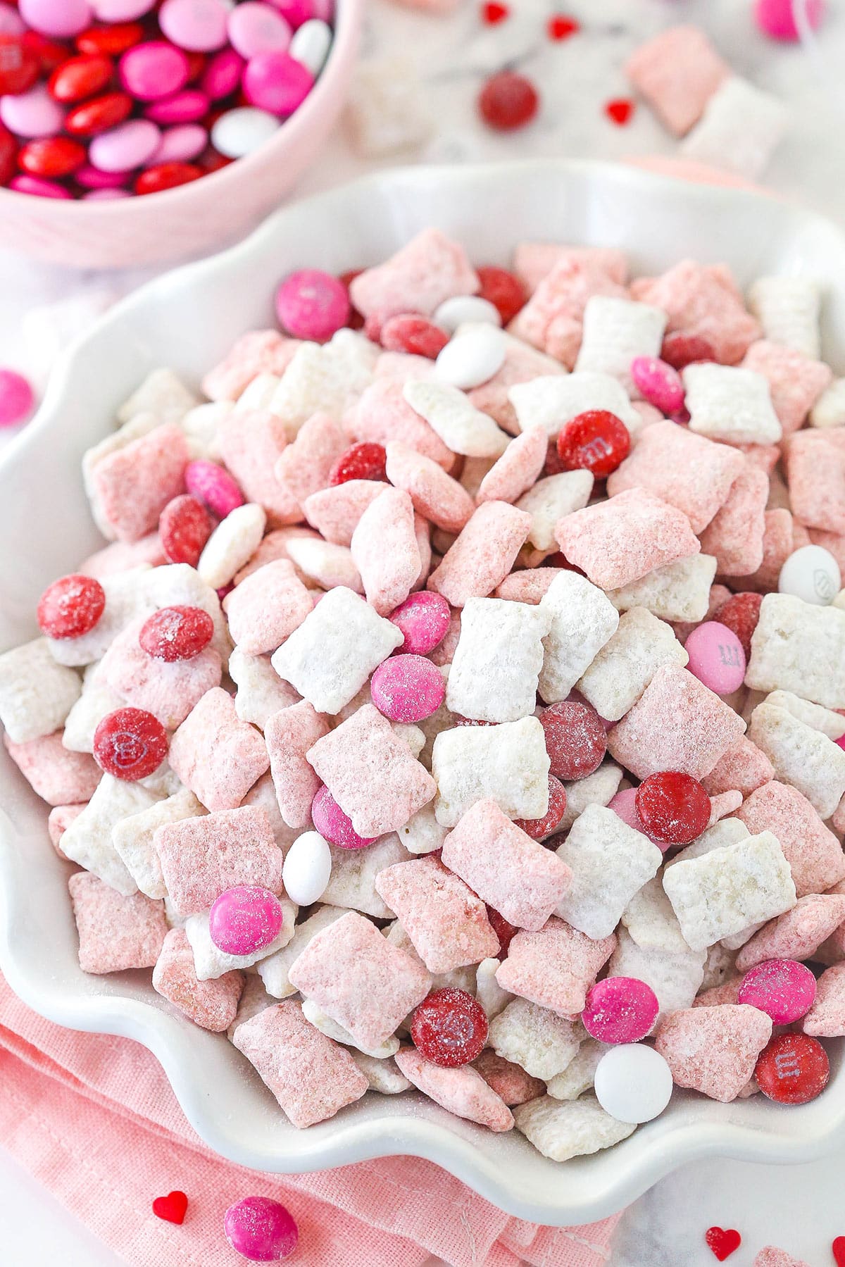 Strawberries and Cream Puppy Chow is perfect for sharing on Valentine's Day! It's an easy, no bake recipe that's loaded with Chex cereal, chocolate, strawberry cake mix and M&Ms! pink and white strawberries and cream puppy chow in white ruffle bowl with m&ms in background