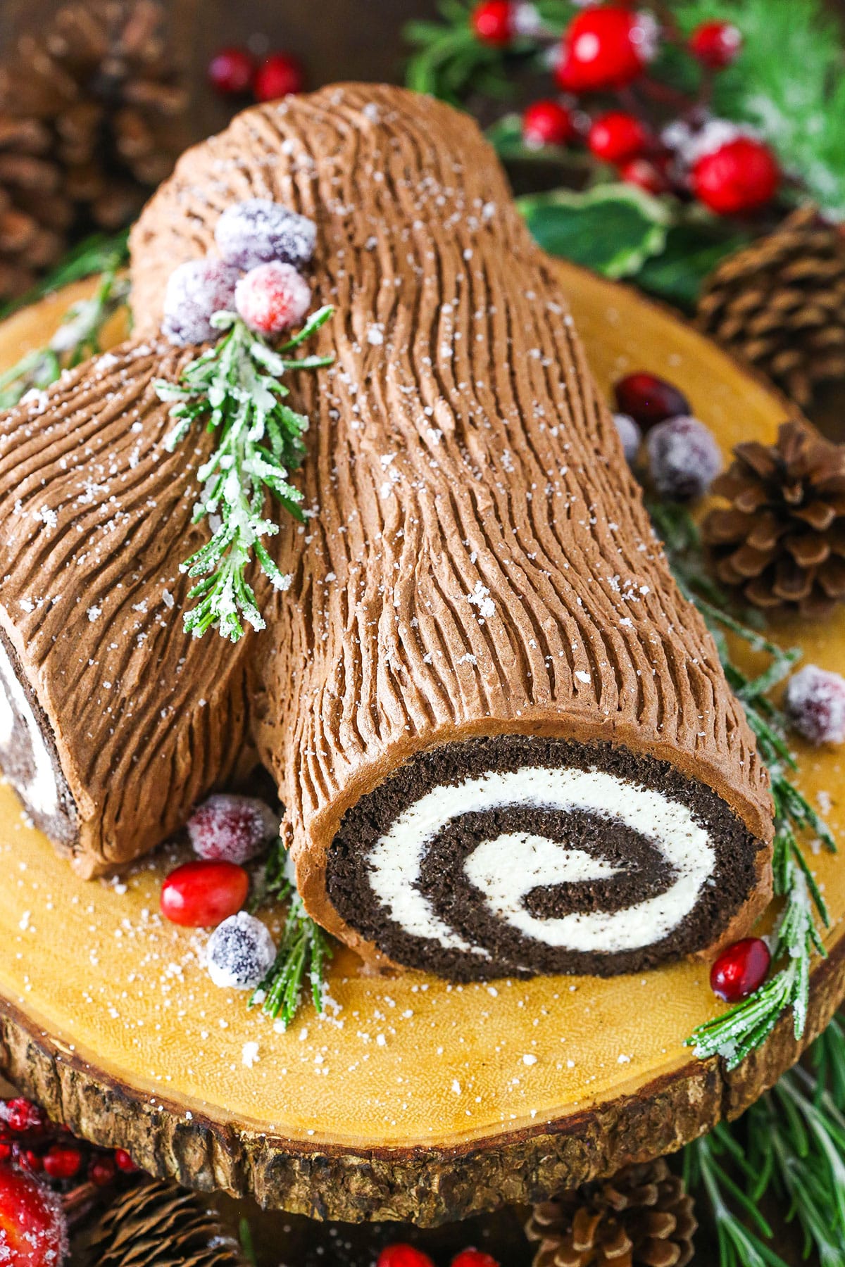 This classic Yule Log Cake is a tender chocolate sponge cake filled with mascarpone whipped cream and covered with whipped chocolate ganache! It's delicious, festive and made completely from scratch! Side view of a Yule Log Cake decorated with sugared cranberries and rosemary sprigs on a wooden cake stand